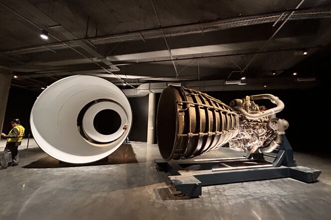 A huge open room with dark floors and walls. A large metal space shuttle engine is displayed towards the right of the image. An even larger stark-white circular solid rocket booster segment is laid on its side to the left.
