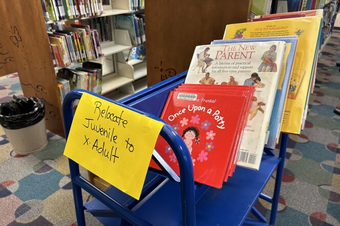 A blue cart in a library with a yellow sign that says "Relocate Juvenile to Adult." On the top shelf of the cart, you can see a red board book titled "Once Upon a Potty," and other books behind it including "The New Parent and "It's So Amazing!"