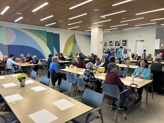 A senior center room filled with at least 7 dining tables with 8 seats each. Older adults are sitting across the tables, some with lunch trays and paper cups in front of them. At least two men can be seen walking the room in aprons and hairnets, handing out food to the older adults.