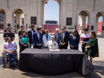 A group of people, including several in USA Olympic and Paralympic team apparel, gather around a table at the entrance arch of the Los Angeles Memorial Coliseum for a document-signing ceremony. A seated official signs papers at the black-draped table while others look on and smile, with the stadium's stands and bronze plaques visible in the background.