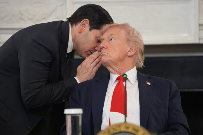 A man leans forward and whispers into the ear of a seated President Trump, who wears a red tie.