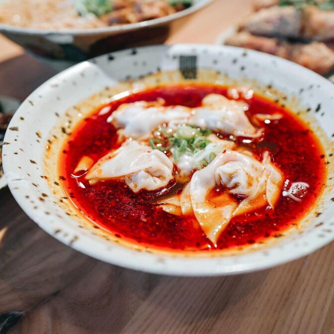 Chinese dumplings in chili oil sauce with scallions on top, served in a speckled bowl.