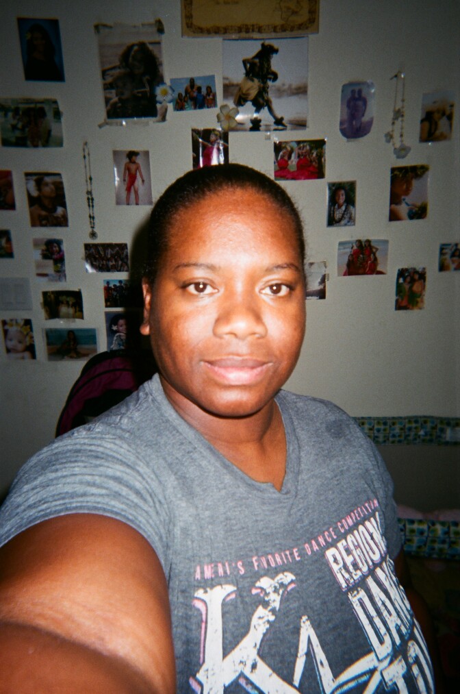 Trina Flowers take a selfie wearing a gray t-shirt with photos taped to the wall behind her.