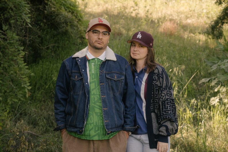 A Latino man with a green soccer jersey, beige pants, a jean jacket, glasses, and an LA baseball cap, stands next to a white woman with a knitted baggy sweater, a dark blue shirt, and a brown LA baseball cap. They are surrounded by grass and greenery.