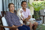 An older woman and man with light brown skin tone sit on chairs on a porch. The woman holds a white chihuahua in her left arm. 