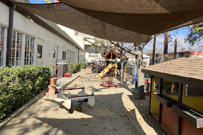 A long playground is next to a one-story white building to its left. The playground has a sand floor, a wooden structure with a slide and various other equipment. To the right is another wooden structure, also on the sand. It's a sunny day and blue sky can be seen at the top of the frame. A green hedge lines the white building.