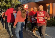 A group of adults walk in a small circle outside a building. They all wear red shirts and carry signs. One visible sign says "Fully staffed schools now" in all capital letters.