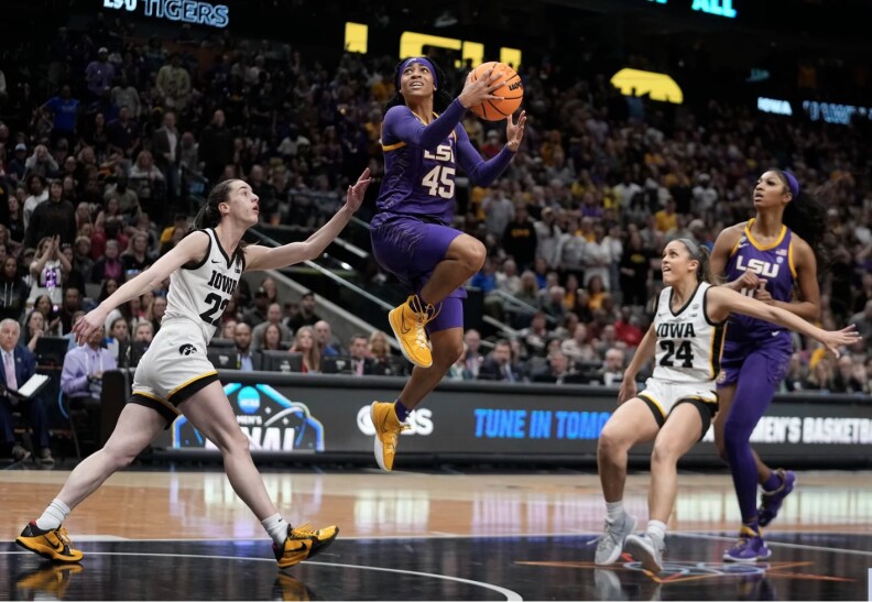 A brown-skinned Black woman leaps in the air to shoot a basketball into a hoop as other players watch.