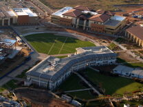 An aerial view of a university campus with a baseball diamond at the center. Several very large buildings encircle the diamond, and a parking lot can be seen in the upper left-hand corner. It is day time. 