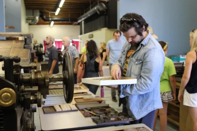 A person examines wooden printing blocks on a table near a vintage letterpress machine as others browse in the background.