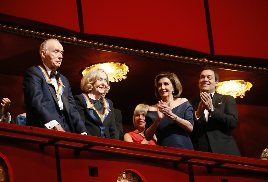 Five people are standing in a red velvet-encased balcony at a theater. One man and one woman, both of whom appear to be in their 70s or 80s, face towards the theater with medals around their necks. Another man and another woman face the first two and applaud.