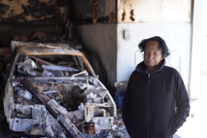 A Black woman stands next to the shell of a burned out car.