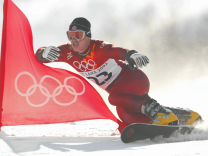 Snowboarder in red olympic suit shreds around a red olympic flag planted in the snow