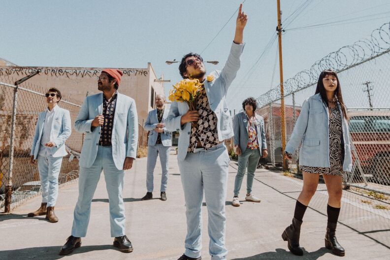 A music group stands in an alley way wearing matching baby blue suits. The man in the center holds yellow flowers.