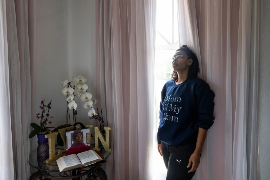 A young Black woman leans against a floor-to-ceiling window inside a home. She gazes up to the left. She wears a sweatrshirt with quilted stitching that reads "Mom of my Mom." The room is relatively dark, with curtains pulled mostly over the windows, letting in just a small amount of light.