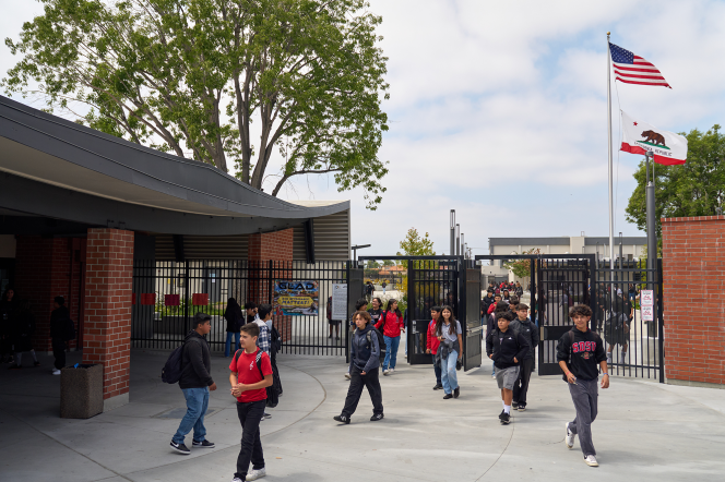 Young kids with backpacks walk through a gate of what looks like a school. It is outdoors and there is a flag pole with an American/California stage flag