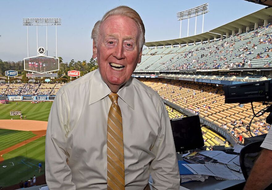 Baseball announcer Vin Scully in a broadcast booth. The Dodger Stadium stands and field are behind him.