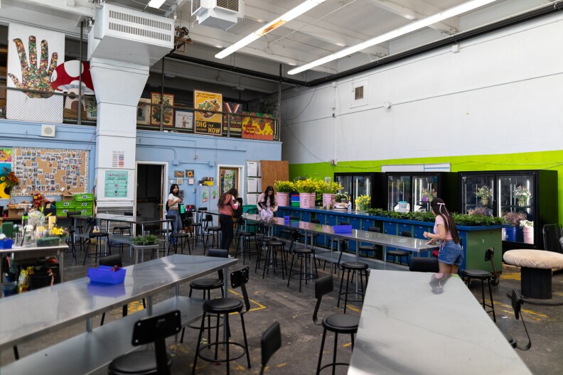 A classroom with long metal tables and black stools. On one wall are various refrigerators with glass doors full of flowers. There are also pink buckets on one table with yellow flowers.