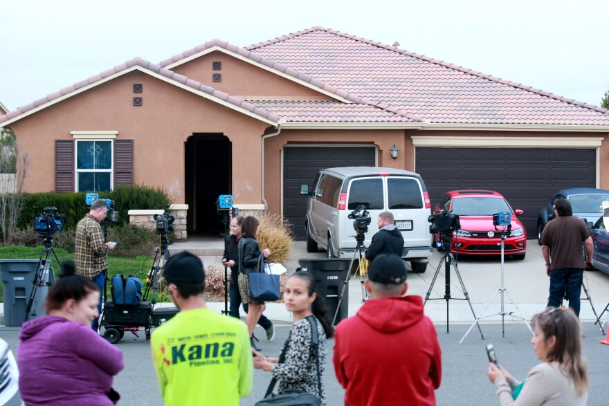 Onlookers and news crews stand in front of a home with brown stucco walls and a tiled roof. Video cameras and lights are propped up on tripods in the street. Several cars are parked in the driveway of the home.