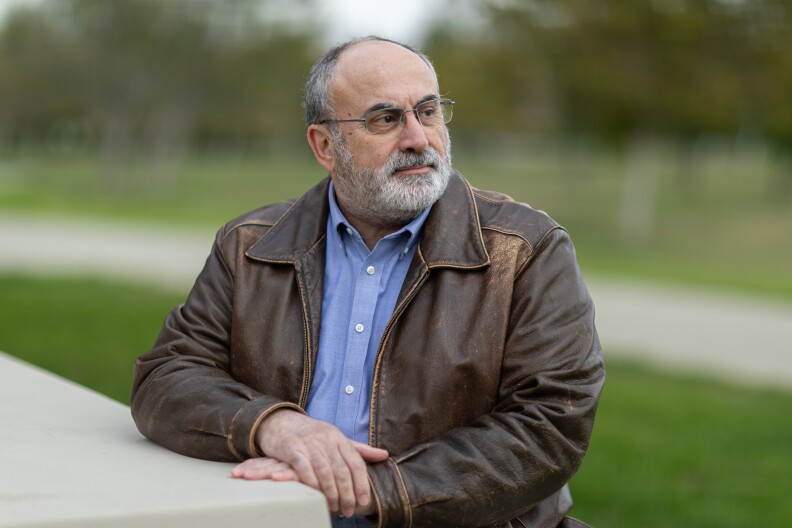 A man with glasses and a gray beard stands in a park, looking off camera to the right, with his hands crossed. He wears a blue shirt and a brown leather jacket.