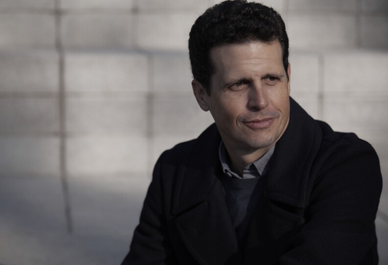 El hombre de piel clara y cabello corto y ondulado viste una chaqueta oscura con un blazer y una camisa con cuello.