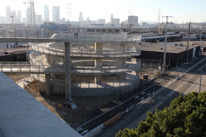 A spiral walkway leads from a bridge down to a street with a two-way designated bike lane running along. Part of the L.A. skyline is seen in the distance.