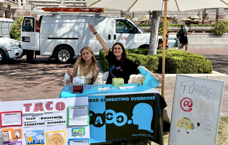 Two young women sit behind a table on a college campus with a signs that say Team Awareness Combatting Overdose