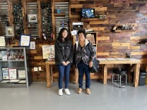 A mother and daughter with dark hair and medium skin stand in front of a wood-covered wall that's covered in award certificates and tech gadgets.