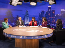 Five people sit at a round table in a TV studio with a Los Angeles skyline backdrop for a debate.