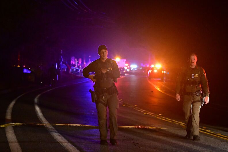 Two people in uniforms and guns holstered on their hips are walking in the middle of a broad rural street at night. Emergency vehicles are in the distance behind them, red and blue lights flashing.