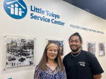 Marian Sunabe and Matthew Yonemura stand side by side inside the Little Tokyo Service Center in Los Angeles. A sign above their head reads "Little Tokyo Service Center: Positive Change for People and Places." They stand in front of a wall of framed photos. The photo to their left is a black and white shot of a street scene from days past. 