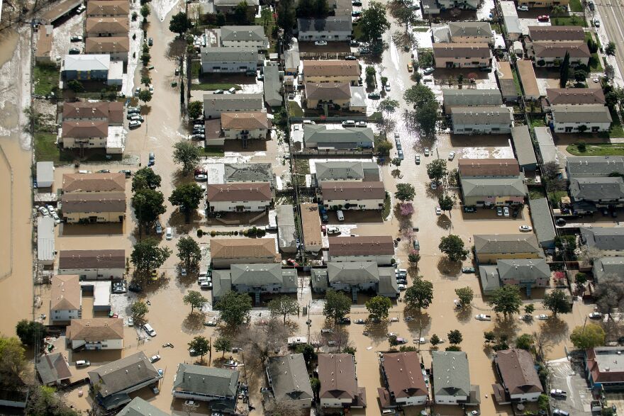 An overhead view from a helicopter showing a neighborhood of two dozen houses with gray, tan, and brown rooves surrounded by light brown flood water.
