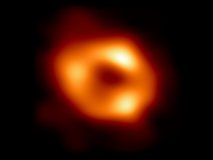 A blurry ring-like image of an event horizon shows an orange glow with three particularly bright areas surrounding a black hole.