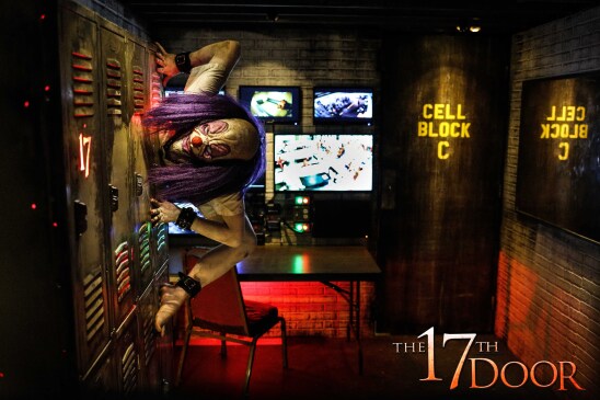 A scary clown with purple hair spider crawls across lockers in a room boasting "Cell Block C" in the right corner. On the back wall are cameras showing other rooms in a penitentiary.