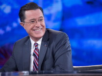 Stephan Colbert, a man with light light skin tone wearing a gray suit, striped tie and glasses, smiles as he looks out of frame to his left.