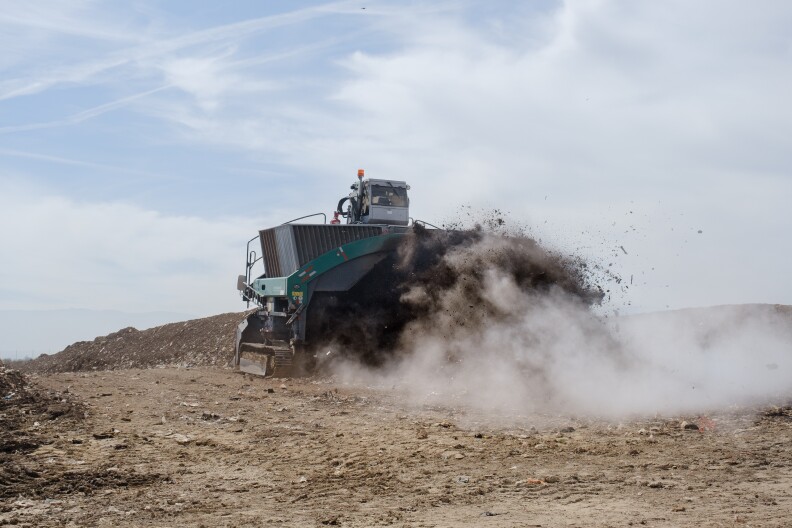 A large machine drives over large rows of compost, kicking up dirt and steam. 