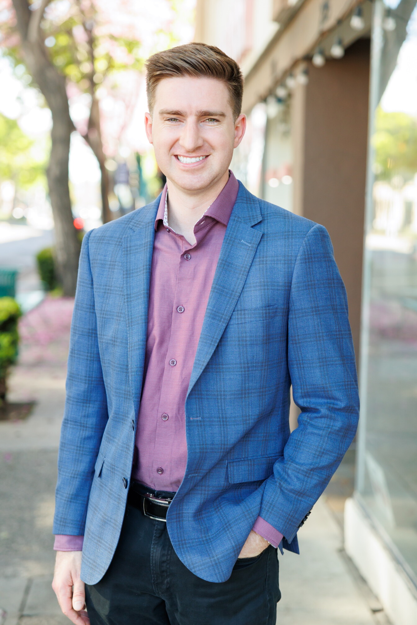 a smiling man with short brown hair wears a blue jacket over a light purple button up collared shirt, and stands outside on the sidewalk