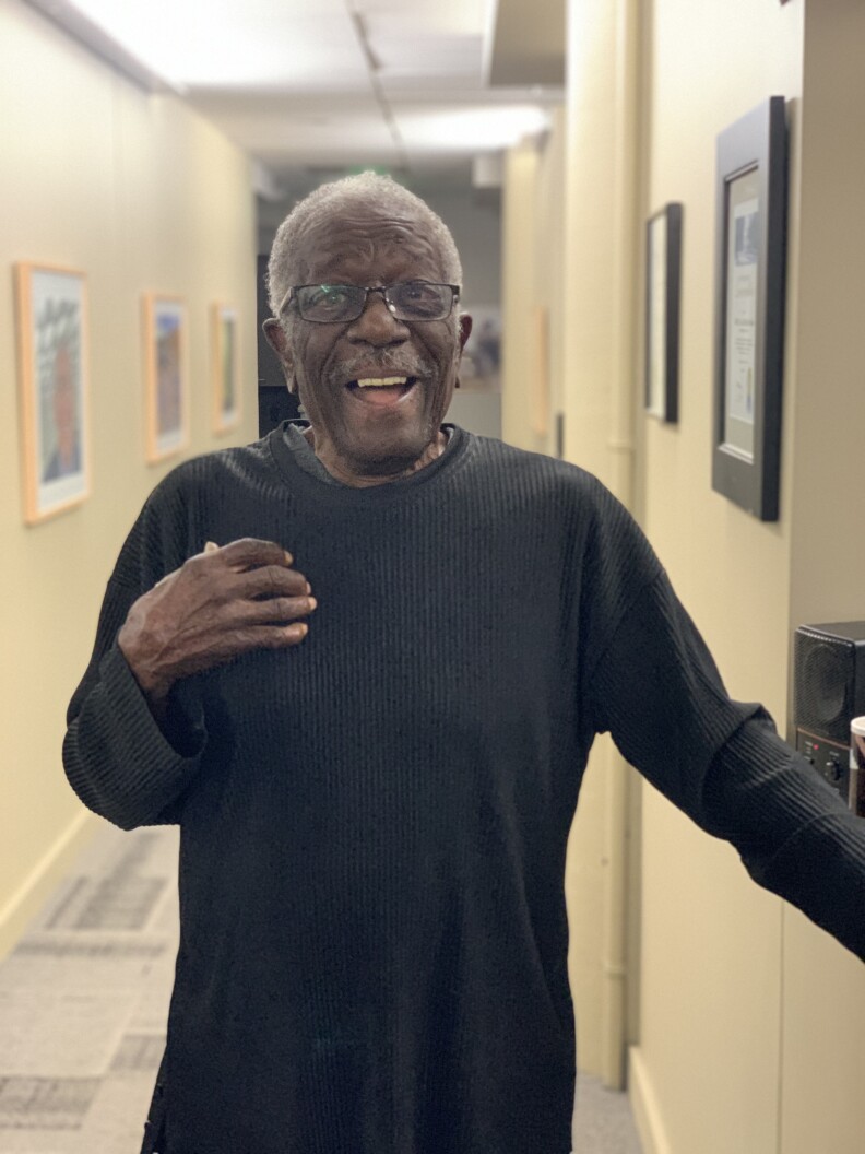 An older Black man with gray hair wears a long-sleeved black shirt and stands in a hallway.