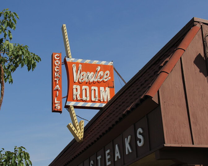 A weathered orange neon sign reading "Cocktails / Venice Room" with an arrow pointing down toward a "Steaks" sign on the building below, set against a blue sky.