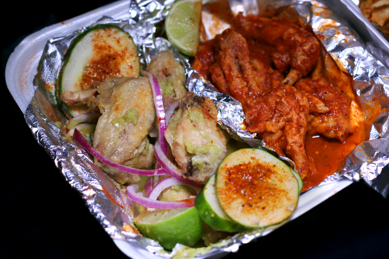 Two kinds of chicken wings sit in foil in a styrofoam container: wings in a pale green sauce on the left and wings in red sauce on the right.