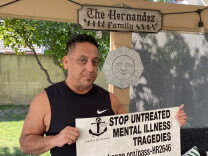 Anthony Hernandez stands in his backyard carrying a banner that reads "Stop Untreated Mental Illness Tragedies." He wears a black tank top. Behind him there is a wooden sign that reads "The Hernandez Family" as well as stone piece of art depicting a sun. 
