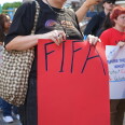 A person with a light skin tone wearing a black t-shirt holds a red poster that reads "FIFA." The image is solely of the person's torso, but behind them you see other demonstrators.