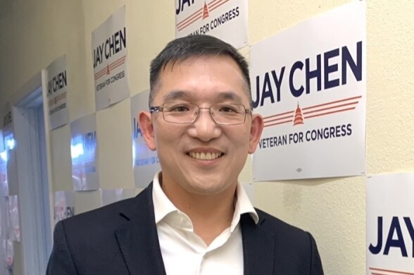 An Asian American man wearing glasses and a jacket poses in a hallway with signs that read "Jay Chen: Veteran for Congress."
