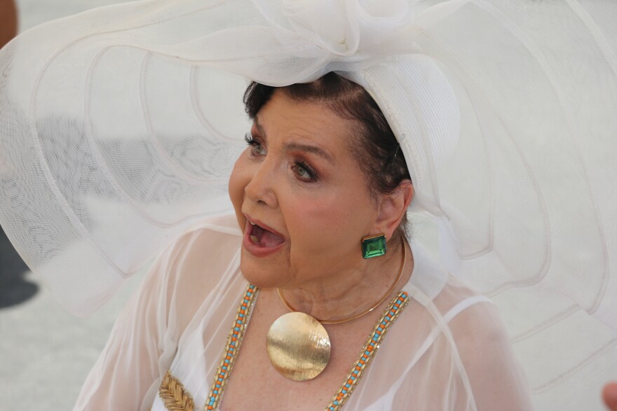 An older woman of mixed heritage wearing a large gold amulet necklace, big green gem earrings and an enormous-brimmed white hat.
