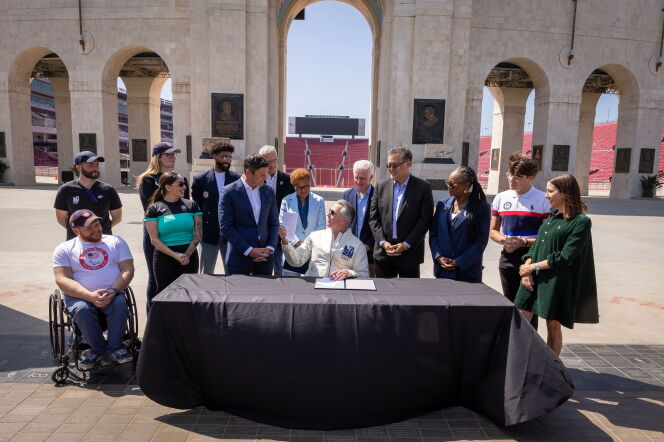 A group of people, including several in USA Olympic and Paralympic team apparel, gather around a table at the entrance arch of the Los Angeles Memorial Coliseum for a document-signing ceremony. A seated official signs papers at the black-draped table while others look on and smile, with the stadium's stands and bronze plaques visible in the background.