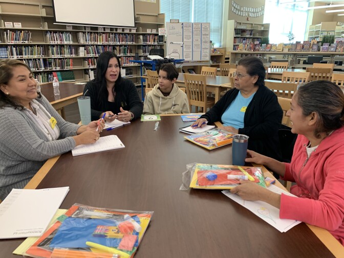 Five women and a boy (center) sit and talk around a table in a room with bookshelves behind them.