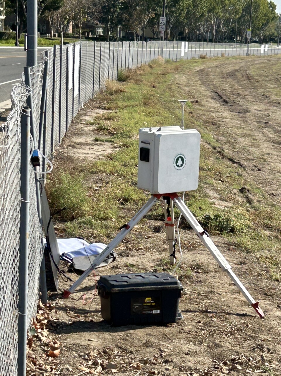 A metal box sits on a tripod attached by wired to a large black box. It sits next to a fenceline. In the distance, you can make out homes and trees across a street on the other side of the fence.  