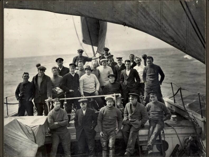 A black and white photo of approximately two dozen young men dressed in sailor's caps and warm sweaters and jackets, standing on the deck of a boat as it sails on the water.