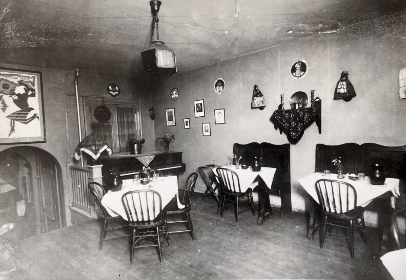A black and white photo of a restaurant's dining room. Three tables are set up with white tablecloths, and a piano sits in the corner.
