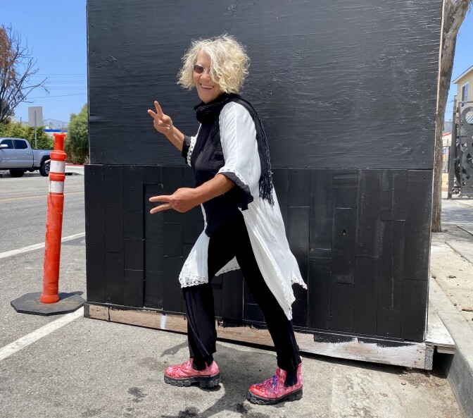 Evans is standing in the street posing and both hands are doing the 'peace' sign. She is smiling and there is a black box behind her. 